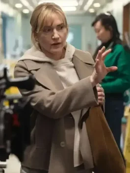 Front view of a woman in a beige coat layered over a light hoodie, holding a brown tote bag and gesturing indoors.