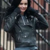 Close-up of Krysten Ritter as Jessica Jones in her black leather biker jacket and fingerless gloves, smiling slightly against a city fence.