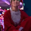 Olivia Holt in a red Santa coat and hat sits with her arms crossed in a brightly lit festive space.