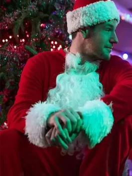 Man in a red Santa coat and hat sitting beside a decorated Christmas tree with holiday lights.
