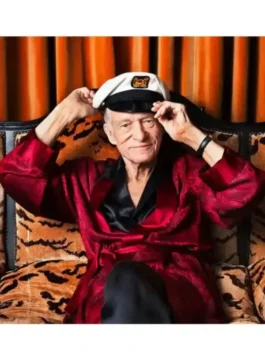 hugh hefner, embodying the hugh hefner costume for men in a red satin robe and black undershirt, seated on a tiger-print couch wearing a captain's cap. Hugh hefner attire for men.