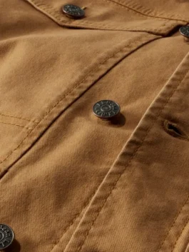 Detailed close-up of a Brown Trucker Jacket showing the durable heavy-duty cotton canvas fabric, reinforced stitching, and branded metal hardware buttons.