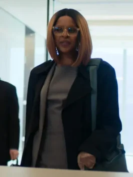Front view of Yusra Warsama in the 2026 series Steal, wearing a structured black trench coat over a grey dress with clear glasses.