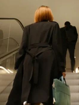 Rear view of the Yusra Warsama black trench coat from the TV series Steal, showcasing the tied fabric waist belt as she rides an escalator.