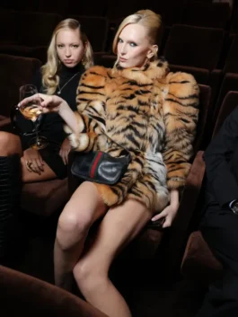 Alex Consani sits in a plush chair wearing a tiger-print fur coat and holding a drink, highlighting the Gucci accessory and glam aesthetic.