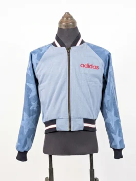 Full-length front view of the zip-up Adidas Denim Graphic Varsity Bomber Jacket featuring a light wash body, red text logo, and star-print sleeves.