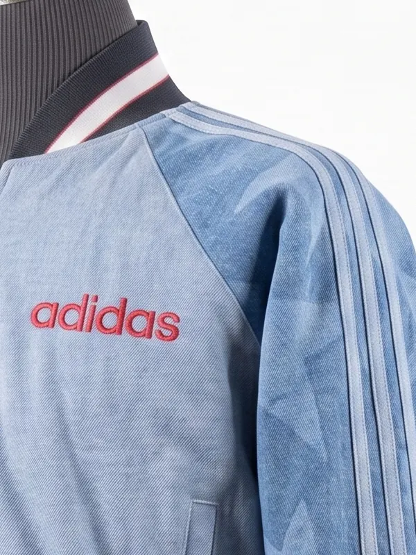 Close-up of the red embroidered Adidas logo on the front upper chest of the light blue denim graphic varsity bomber jacket.