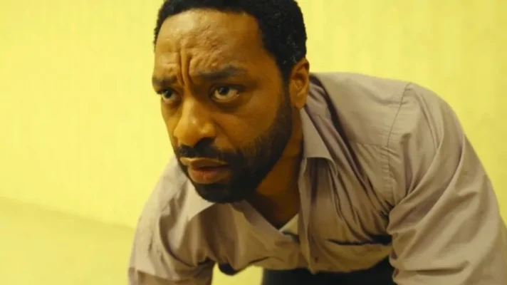 Chiwetel Ejiofor as Clark in the Backrooms movie 2026, leaning forward with fear against the yellow liminal space wallpaper
