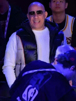 Vin Diesel smiling in a white crewneck shirt and a black and grey hooded puffer vest during a basketball event.