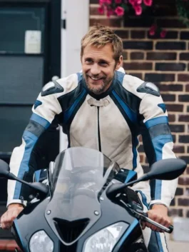 Alexander Skarsgard smiling while riding a black sport motorcycle in a white, blue, and black leather biker jacket