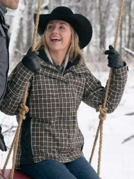 Candid view of Amy Fleming smiling on a wooden swing in a snowy forest wearing the brown plaid Heartland winter coat.