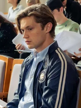 Seated profile view of Damian Hardung as James Beaufort in Maxton Hall wearing a dark blue bomber jacket with collar snap detailing and chest patch.
