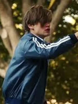 Side action shot of Damian Hardung as James Beaufort wearing a blue track bomber jacket with white sleeve stripes.