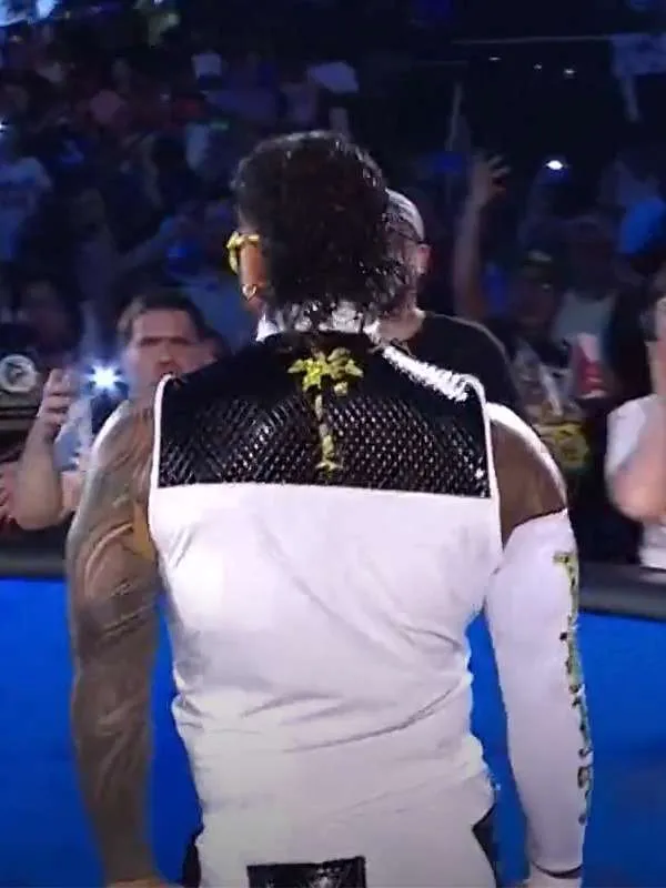 Rear view of WWE wrestler Jey Uso walking through a crowd, wearing a white sleeveless top with a textured black upper panel featuring a gold palm tree design on the back.