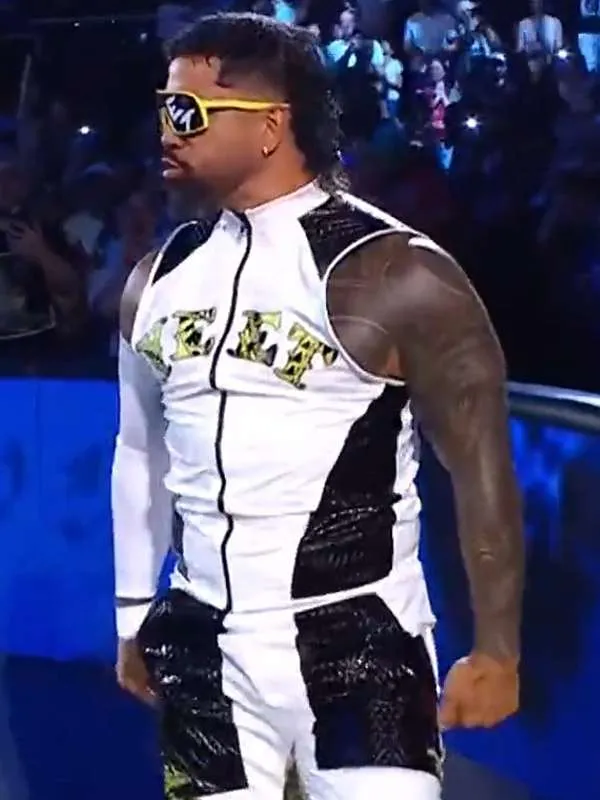 Side-angle profile shot of Jey Uso standing in the ring, showing his tattooed arm, slicked-back hair with a mullet, yellow sunglasses, and his white and black zippered wrestling vest.