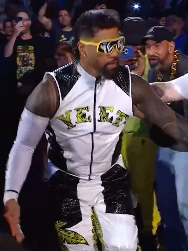 Jey Uso walking past fans and crew, wearing yellow-framed "YEET" sunglasses and his white and black sleeveless wrestling vest with yellow "YEET" lettering.