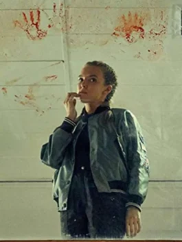 Low-angle mirror reflection shot of Jodie Comer as Villanelle wearing a silver satin bomber jacket with striped rib-knit trim, sleeve patches, and snap-button front.