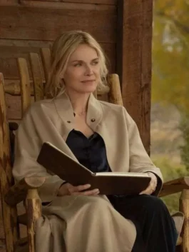 Close-up of Michelle Pfeiffer sitting on a wooden chair reading a book, wearing a beige wool trench coat with a button collar.