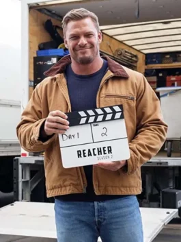 Alan Ritchson smiling on set behind the scenes of Reacher Season 2 holding a film slate board, wearing a brown corduroy-collar cotton jacket.