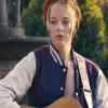 Front view of Ellie de Lange wearing a purple and white varsity bomber jacket with silver snap-button closures while playing an acoustic guitar.