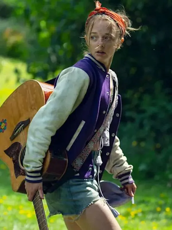Side profile of Ellie de Lange walking through a grassy field wearing the purple and cream varsity jacket while carrying an acoustic guitar over her shoulder.