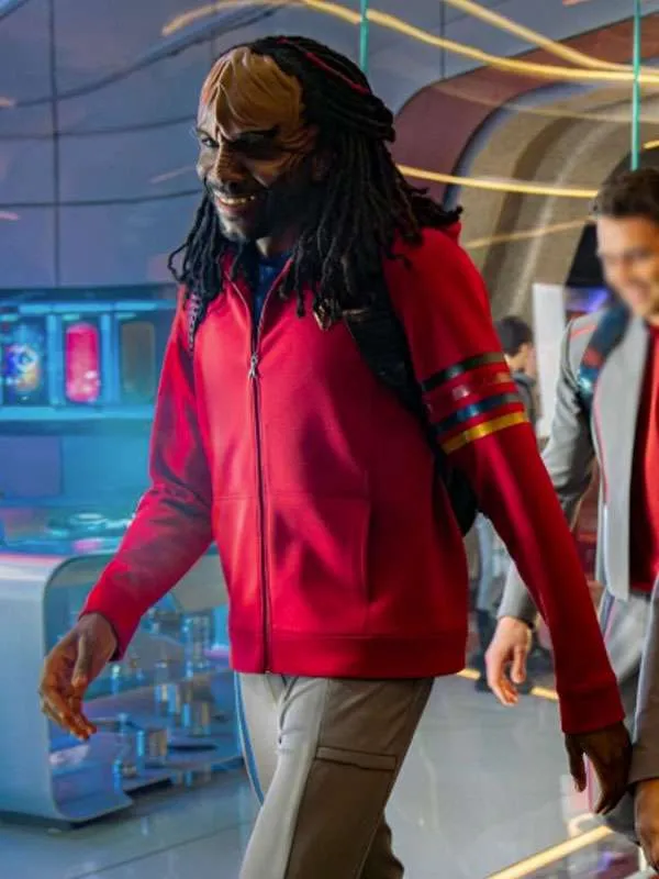 Karim Diane as a cadet in Star Trek: Starfleet Academy, wearing a casual red zip-up hoodie with multi-colored rank stripes on the sleeve while seated in a futuristic lounge.