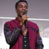A young man holding a microphone while wearing a maroon Starfleet Academy varsity letterman jacket with black leather sleeves and "SFA" chest embroidery.
