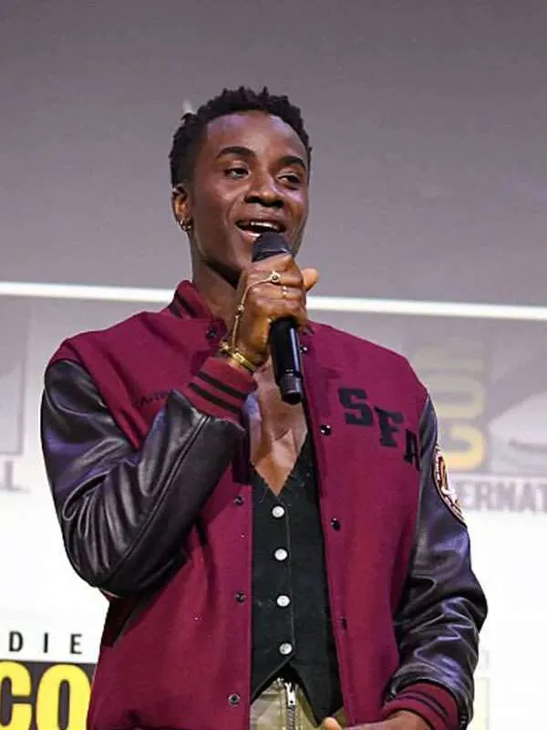 A young man holding a microphone while wearing a maroon Starfleet Academy varsity letterman jacket with black leather sleeves and "SFA" chest embroidery.