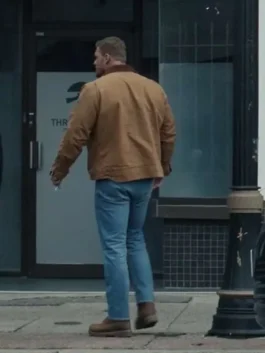 Back view of Alan Ritchson as Jack Reacher walking down a sidewalk wearing a brown utility jacket and blue jeans.
