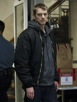 Joel Kinnaman as Stephen Holder standing indoors wearing a black bomber jacket layered with a grey zip-up hoodie.