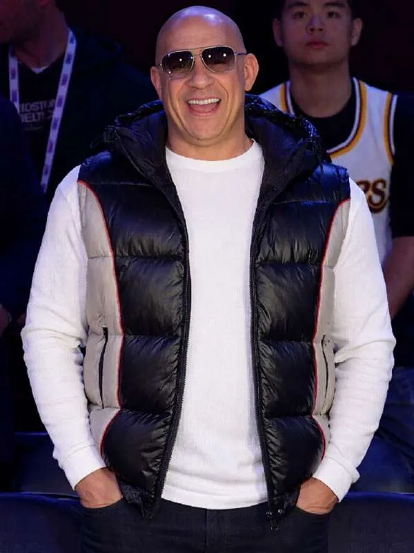 Front view of Vin Diesel standing with hands in his pockets, wearing his black and grey 75th All-Star Game hooded puffer vest and sunglasses.