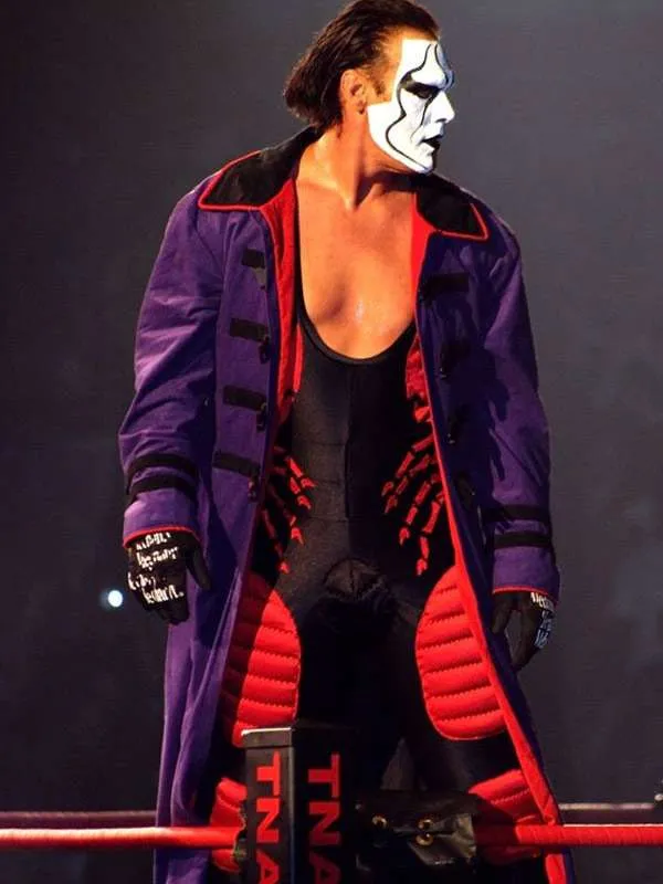Three-quarter shot of wrestler Sting standing in a dark arena, wearing his long purple trench coat over a black singlet with red skeleton hand prints.